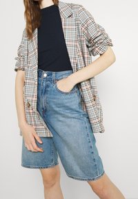 Blazer a quadri nei colori pastello sopra una maglietta a coste blu navy, abbinato a pantaloni shorts in denim a vita alta di un blu chiaro. Presenta una silhouette aderente.