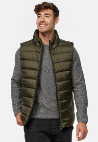 Oliven grøn puffer vest med høj krave og lynlås, præget af en quiltet tekstur. Båret over en grå sweater, med sorte bukser.