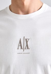 Hvid bomulds t-shirt med et forhøjet beige broderet logo "A/X" og teksten "ARMANI EXCHANGE" nedenunder, rund halsudskæring og korte ærmer.