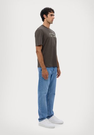 T-shirt in cotone grigio scuro con grafica frontale, abbinato a jeans azzurri e sneakers bianche. Modello in posizione di profilo su uno sfondo neutro.