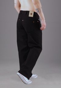 Schwarze Jeanshose mit lockerer Passform, ausgestattet mit einem Patch-Label am hinteren Bund, Kupferriegeln und hochgekrempelten Säumen über weißen Turnschuhen.
