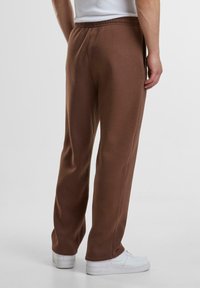 Brune athletic bukser med en afslappet pasform, elastisk talje og sidelommer, parret med hvide sneakers på en neutral baggrund.
