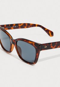 Sluneční brýle s želvovinovým vzorem, tmavými obdélníkovými skly a robustními ramínky, nakloněné na bílém povrchu.