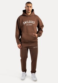 Hoodie marron et jogging assorti avec le texte blanc "SMILODOX ATHL WEAR". Le tissu semble doux, avec une coupe décontractée et une poche avant. Baskets blanches.