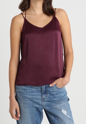 Woman wearing a sleeveless, dark purple satin camisole top paired with light blue distressed jeans, hand in pocket, against plain background.