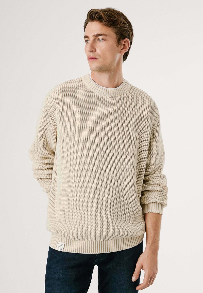 Young man wearing a beige knitted sweater and dark jeans, looking to the side with hands relaxed at waist level against a plain background.