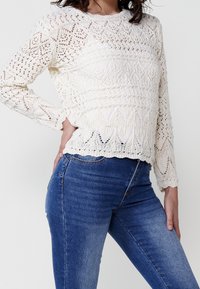 Krémově zbarvený pletený pulovr s propracovanými krajkovými vzory a žebrovaným lemem. Má dlouhé rukávy a kulatý výstřih, doplněný modrými džínami.