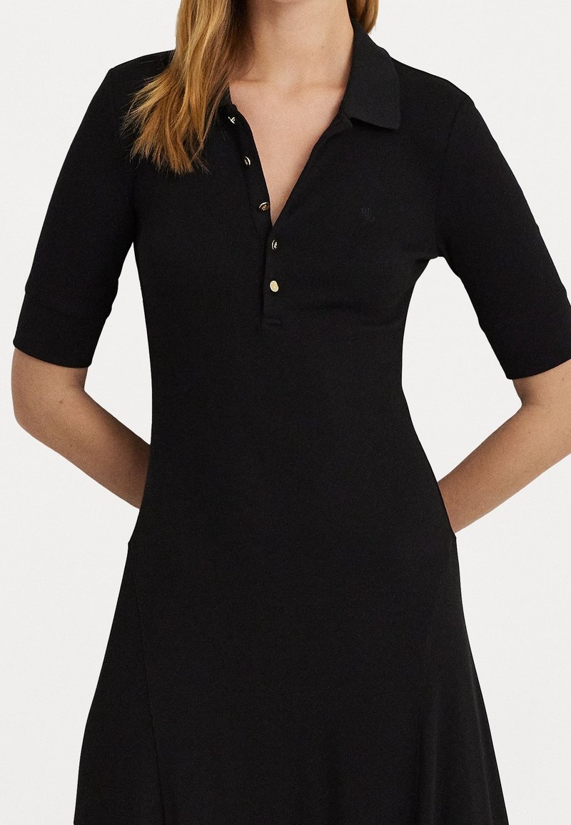 Robe polo noire à manches courtes, dotée d'un col à boutons et d'accents en métal doré, confectionnée dans un tissu lisse et texturé.