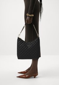 Borsa a tracolla in pelle nera trapuntata con forma curvada, manico in catena argentata e emblema triangolare sul davanti. Tacchi a punta in vernice marrone.