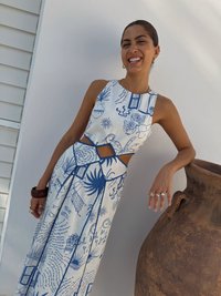 Rochie albă cu modele albastre, având plante și text. Decupaj la talie, design plisat, asociată cu o vază din ceramică maro.
