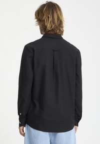 Chemise noire à manches longues en tissu lisse, avec un pli au dos, des poignets à boutons et un col classique. Design à ourlet arrondi.