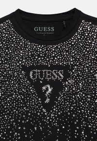 Μαύρο κοντομάνικο μπλουζάκι με σχέδιο από διάφανα στρας. Το κεντρικό γραφικό περιλαμβάνει το όνομα της μάρκας "GUESS" και ένα ερωτηματικό.