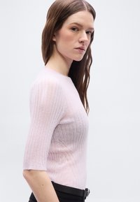 Νεαρή γυναίκα με μακριά καστανά μαλλιά που φοράει ροζ ριμπ πουλόβερ και μαύρο παντελόνι, στέκεται μπροστά σε απλό φωτεινό φόντο.