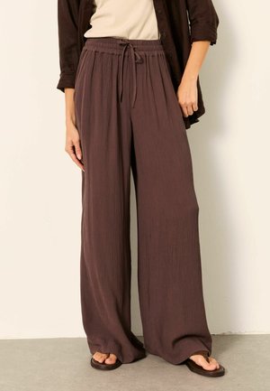 Woman wearing loose, brown textured wide-leg pants with an elastic drawstring waist, paired with brown flip-flops and a beige top.