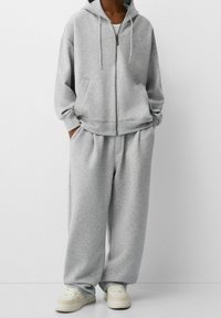 Persona con sudadera gris holgada con cremallera, pantalones deportivos anchos a juego, camiseta blanca y zapatillas blancas, de pie frente a un fondo liso.