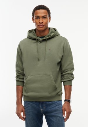 Hombre con sudadera con capucha verde oliva con bolsillo delantero y vaqueros azules, de pie frente a un fondo blanco liso.