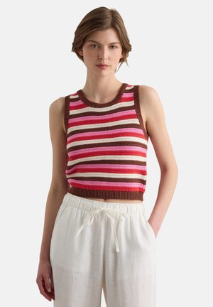 Frau trägt ärmelloses, gestricktes Croptop mit horizontalen braunen, pinken und roten Streifen, kombiniert mit lockeren weißen Hosen mit Kordelzug.