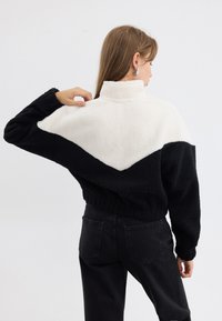 Giacca in pile bianca e nera con colletto alto, caratterizzata da un design a zig zag. Il tessuto sembra morbido e testurizzato, con polsini e orlo elasticizzati.