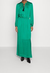 Grön långärmad klänning i silkig kontakt, med rund halsringning, dragsko med tofs och en avslappnad silhuett.