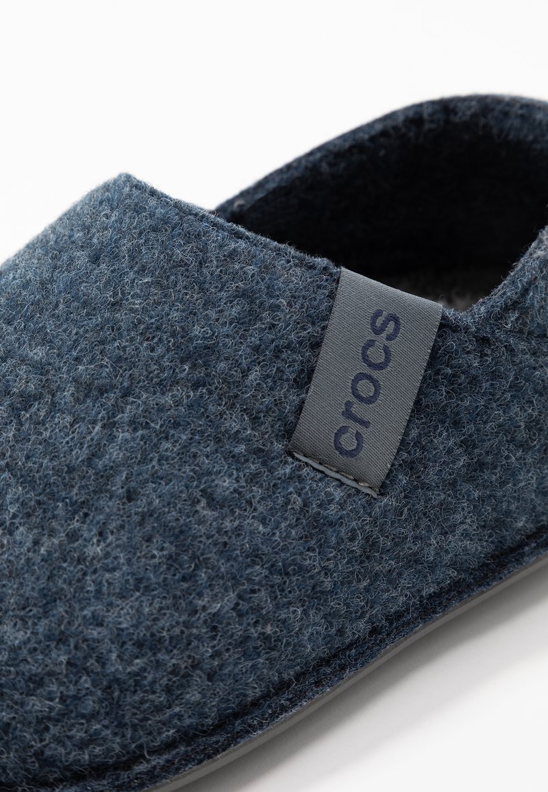 Zapatilla de fieltro azul marino con una etiqueta gris de Crocs, forma redondeada y superficie texturizada, con un suave forro interior y suela de goma.
