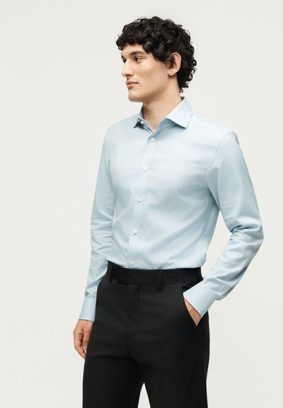 Νέος άνδρας με σγουρά μαλλιά που φοράει ανοιχτά μπλε πουκάμισο με κουμπιά και μαύρο παντελόνι, στέκεται με το ένα χέρι στην τσέπη, κοιτάζοντας στην πλευρά.