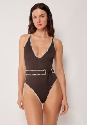 Frau mit langen dunklen Haaren, die einen strukturierten dunkelbraunen Einteiler mit weißen Absetzungen und Gürtel trägt, vor einem schlichten Hintergrund stehend.