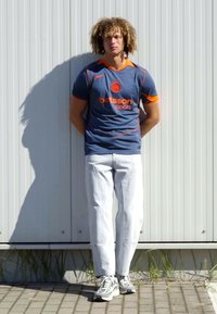 Maillot de sport bleu avec des accents orange, jeans blancs et baskets grises, se tenant contre un mur gris clair. Ombre visible sur le sol.