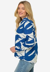 Blusa a maniche lunghe blu e bianca con bold patterns astratti, colletto alto e polsini elastici, abbinata a jeans azzurri chiari.