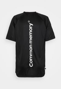 Jersey deportivo negro con cuello redondo y mangas cortas. Presenta un texto vertical en blanco que dice "memoria común" en la parte de atrás. Textura suave.