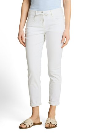 Kobieta ubrana w podwinięte białe dżinsy, beżowe klapki i jasnoniebieską bluzkę, stojąca z rękami swobodnie opuszczonymi wzdłuż ciała.