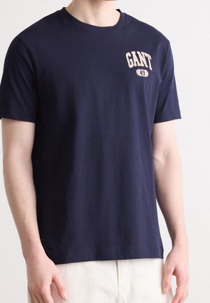Mężczyzna noszący granatowy t-shirt z krótkim rękawem z napisem „GANT 49” w beżowym kolorze na górnej lewej części klatki piersiowej.