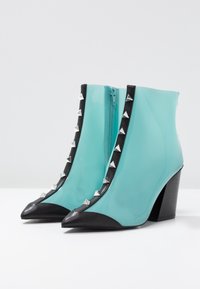 Bottines turquoise avec des bouts pointus noirs, des talons épais et des bordures noires ornées de clous en pyramide argentés et d'une fermeture éclair sur le côté.
