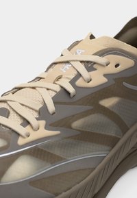 Κοντινό πλάνο αθλητικού παπουτσιού σε μπεζ και γκρι χρώμα με πάνω μέρος από δίχτυ, αφηρημένες επικαλύψεις και μπεζ κορδόνια δεμένα πάνω από τη γλώσσα.