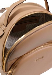 Rund ryggsäck i ljusbrunt läder med två dragkedjeförsedda fack, guldfärgade beslag och ett mönstrat innerfoder med en subtil logotyp.