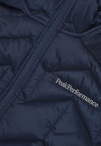 Sötétkék pehelykabát átlós tűzéssel, cipzáros záródással és ezüst hímzésű "PeakPerformance" logóval. Sima, fényes felület.