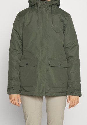 Person iført en olivengrøn hættetrøje vinterjakke med lommer med klap foran og beige bukser, stående mod en ensfarvet baggrund.