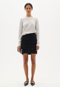 Svetlo šedý sveter s dlhým rukávom v kombinácii s čiernou tweedovou minisukňou a čiernymi topánkami s ostrou špičkou, drží čierny clutch.