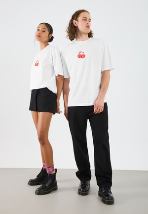Due persone indossano t-shirt bianche con una grafica di ciliegia rossa. La donna abbina la sua a pantaloni corti neri e stivali neri; l'uomo indossa pantaloni neri.