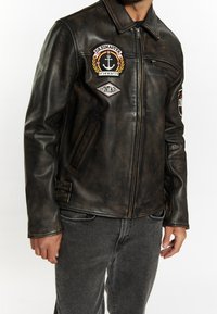 Ruskea nahkatakki kuluneella viimeistelyllä, jossa on kirjailtuja merkkejä, vetoketjullinen suljenta ja sivutaskut. Koristeellisina yksityiskohtina koristenaulat.