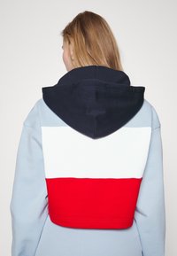 Huvtröja med blå kropp, vit horisontell rand och röd nederdel. Har en marinblå huva och slät textur.