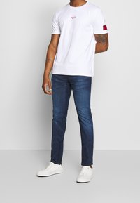 Camiseta de algodón blanca con un pequeño logo rojo en el pecho, combinada con jeans de mezclilla azul oscuro y zapatillas blancas. Diseño minimalista.