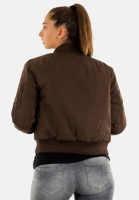Donna con coda di cavallo castana, che indossa una giacca bomber marrone scuro e jeans grigi, rivolta di lato su uno sfondo bianco semplice.
