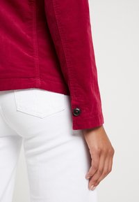 Oikea käsi punaisessa pitkähihhain-paidassa, jossa musta nappi käsivarren lähellä, valkoisten housujen taskun vieressä vaalealla taustalla.