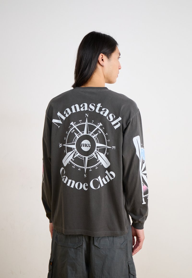 Long-sleeve dark gray t-shirt featuring a compass graphic and "Manastash Canoe Club" text. Additional graphics on sleeves in pastel colors.