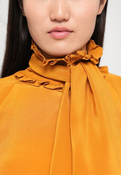 Mosterdgele blouse met een gerimpelde hoge kraag en strikdetails. Gladde, glanzende stof met een subtiele glans en opmerkelijke gathered accenten.