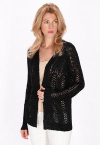 Cardigan nero in maglia con un design in pizzo, apertura frontale, maniche lunghe e polsini a costine. Indossato sopra un top beige e abbinato a pantaloni bianchi.