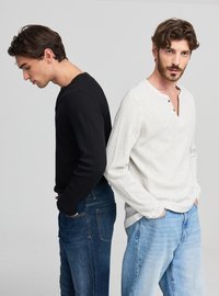 Due uomini stanno schiena contro schiena, uno con una henley nera a coste e maniche lunghe e l'altro con una henley grigio chiaro con patta con bottoni. Entrambi indossano jeans.