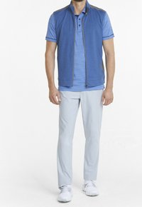 Blå zip-up väst över en ljusblå kortärmad polo, matchad med ljusgrå byxor och vita sneakers. Västen har grå detaljer.