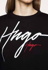 Černé tričko s kulatým výstřihem a velkým bílým a menším červeným nápisem "Hugo" napsaným rukou přes hruď, které nosí osoba s dlouhými hnědými vlasy.