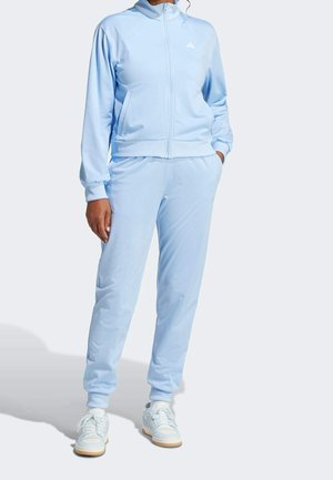 Osoba w jasnoniebieskim dresie na zamek i białych sneakersach, stojąca z jedną ręką w kieszeni na jednolitym tle.